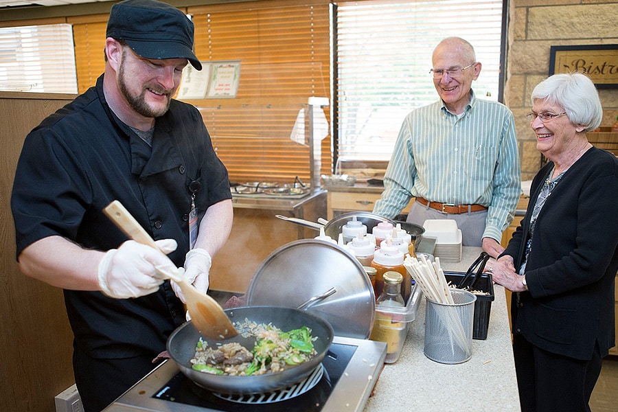 Male Chef Cooking In Pan With Two Residents Watching